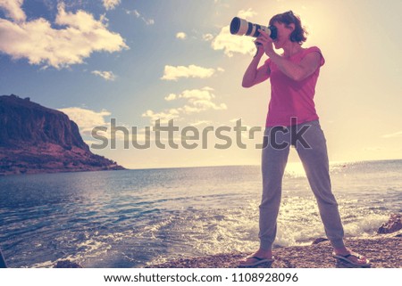 Woman with camera taking photo on seacoast. Rocky island Monemvasia in the background. Greece, Peloponnese, Lakonia. Travel destinations.