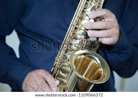A Caucasian male wearing a blue shirt holding and playing a gold platted tenor saxophone against a white backdrop 