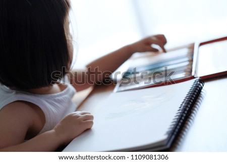 Young Japanese girl drawing at home on a white notebook, sitting at a wooden desk, in a bright room.