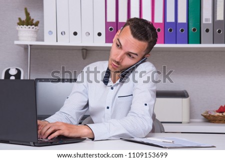 Portrait of a handsome young male manager in an office sitting at a table with a laptop talking on the phone. He sits right in front of the camera smiling and looks serious