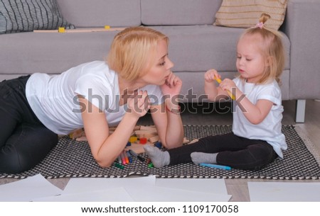 Little charming blond girl painting felt-pen with her mom sitting near the sofa