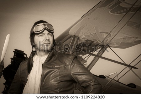 Portrait of a vintage pilot with leather cap, scarf and aviator glasses in front of a historic airplane biplane - Portrait of a man in historical pilot clothing - vintage old picture style
