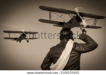 
Portrait of a vintage pilot with leather cap, scarf and aviator glasses in front of a flying historical biplane - Portrait of a man in historical pilot clothing - vintage old picture style
