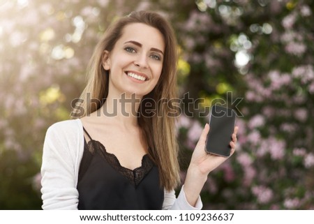 smiling long-haired girl on the background of green foliage of trees holding a smartphone with an empty display.
