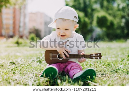 A small child in a cap sits on the grass and plays the guitar.