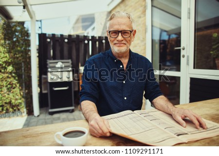 Senior man smiling while sitting at his patio table reading the financial pages of a newspaper and drinking a coffee