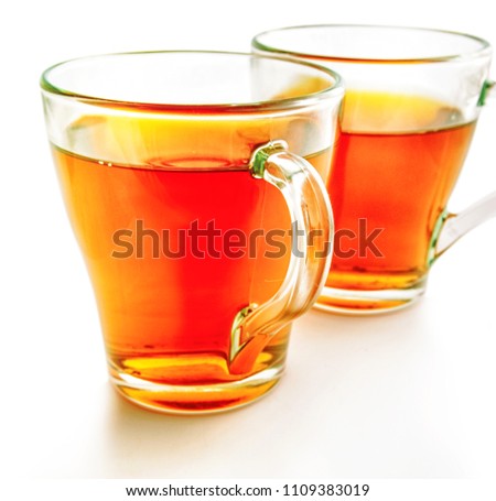 Two glass cups of tea on a white background. In the foreground a sharpness zone on the cup handle. Other image in the small depth of sharpness. Indoors. Color. Photo.