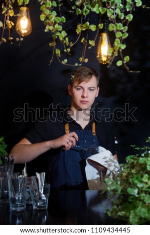 A handsome man cleans a wine glass in a bar on a black background with flashlights. Happy male barman or waiter in apron wiping the fouger.