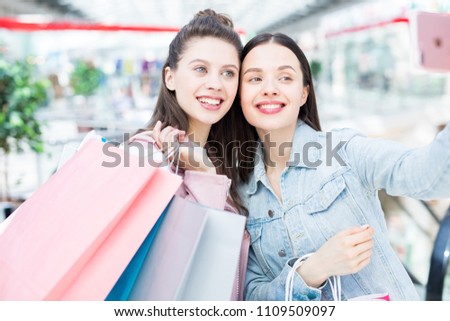Smiling attractive young women in casual clothing photographing together while having fun in shopping mall and buying clothes