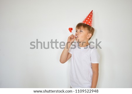 Portrait of cheerful Caucasian boy wearing holiday cap and casual white t-shirt blowing whistle, having fun while celebrating his 10th birthday, standing against blank studio wall background