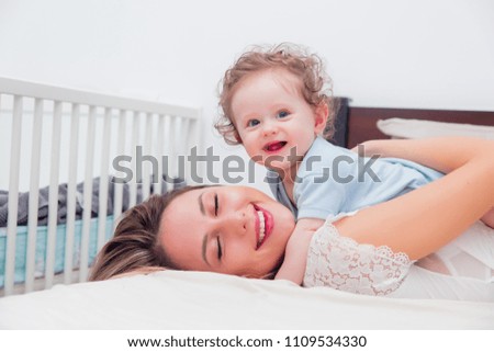 Young mother with her 7 month old little son dressed in pajamas are relaxing and playing in the bed at the weekend together, lazy morning, warm and cozy scene.