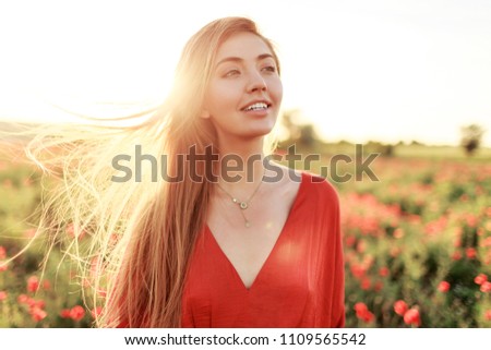 Gentle blonde long-haired woman with perfect smile posing on poppy field in warm summer sunset.  Windy hairs. 