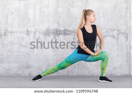 Healthy sportswoman stretches legs wears print leggings, sneakers, prepares for sport competiton, trains body and warms up muscles, poses against grey concrete wall. Woman gets ready to cardio running