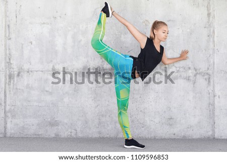Fitness and healthy lifestyle concept. Flexible slim sportswoman prepares for marathon, has exhausting training, raises legs, shows horizontal stand, wears colourful leggings, stands against grey wall