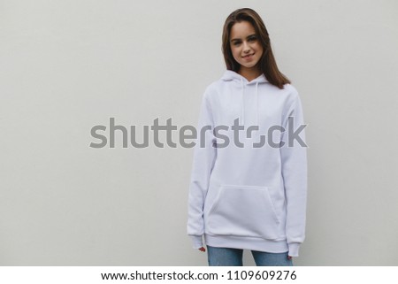 Young pretty girl wearing blank white sweatshirt with space for your logo or design, mock-up of white cotton hoodie, white wall in the background. Front view