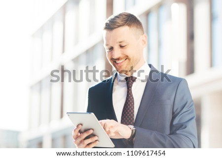 Outdoor shot of prosperous delighted male entrepreneur searches successful ideas for business in internet, uses modern tablet computer, dressed in formal suit. Male economist with digital device