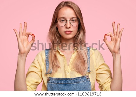 Portrait of attractive serious female with European appearance, wears round glasses, makes okay sign, dressed in casual outfit, isolated over pink background. People and body language concept