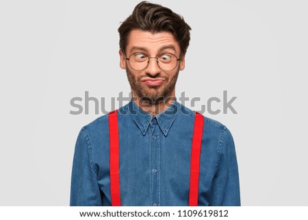 Comic bearded funny young male employee has fun, crosses eyes and makes grimace, acts like little naughty child, wears fashionable denim shirt with red braces, isolated over white background