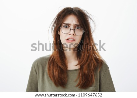 Portrait of untidy funny nerdy woman in trendy transparent glasses with messy hair, looking confused and unfocused, frowning while looking at lots of work to do, unwilling and wanting rest