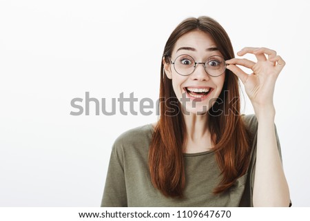 Attractive nerdy girl fascinated to see new book in store. Good-looking overwhelmed woman in casual t-shirt, smiling broadly, seeing something surprising and wonderful, holding hand on rim of glasses