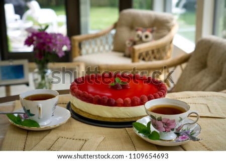 Teatime on a summer porch with beige wicker furniture. Raspberry cheesecake and two cups of black tea on hand-woven straw place mats.