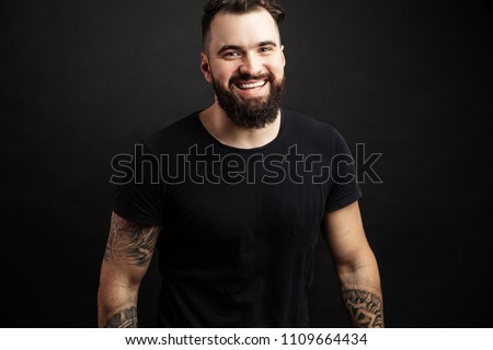 Handsome young biker with tattooed hands in casual black wear smiling while standing against black background. Positive Emotions. Sthength, Power, Humanity and People Concept. Royalty-Free Stock Photo #1109664434
