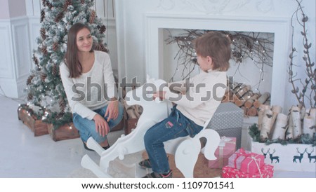 Little boy riding wooden rocking horse while his mom sitting next to Christmas fireplace