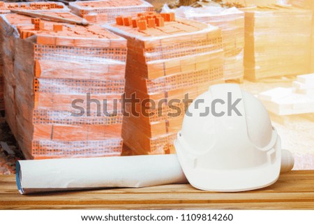 white safety helmet plastic, paper roll plan blueprint on wood floor table and pile brick orange background