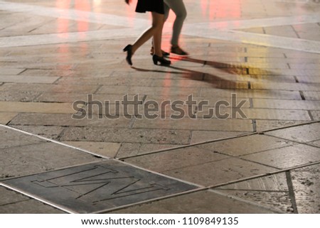 Close up outdoor view of feet and legs of people walking in an urban place during the evening. Silhouette of persons with a bright pink surface in background. Abstract image of urban night activity.