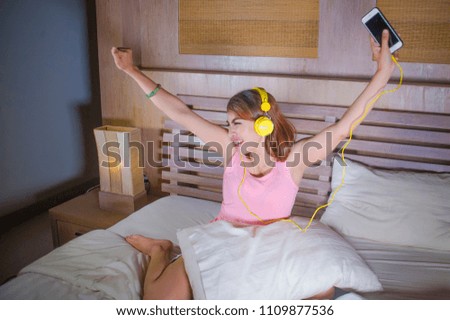 young attractive and happy girl in bed with yellow headphones listening to music on internet with mobile phone dancing and singing crazy excited at home on her bedroom in lifestyle concept