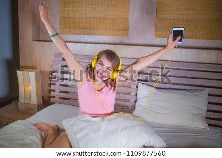 young attractive and happy woman in bed with yellow headphones listening to music on internet with mobile phone dancing and singing crazy excited at home on her bedroom in lifestyle concept
