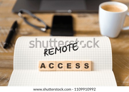Closeup on notebook over vintage desk surface, front focus on wooden blocks with letters making Remote Access text. Business concept image with office tools and coffee cup in background