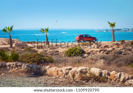 One red hatchback car among short palm trees, green bushes, and stones on the background of blue sea surface. Concept of adventure and traveling, happy vacation