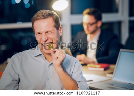 Funny mature man bites shiny golden bitcoin. Middle aged man wearing blue coloured shirt angrily biting golden bitcoin coin with his coworker working in background. Royalty-Free Stock Photo #1109921117