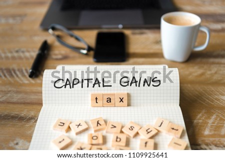 Closeup on notebook over vintage desk surface, front focus on wooden blocks with letters making Capital Gains Tax text. Business concept image with office tools and coffee cup in background Royalty-Free Stock Photo #1109925641