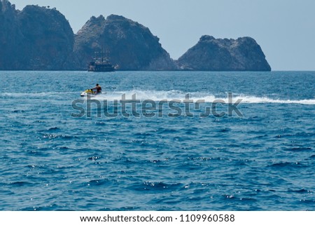              Men in lifejacket riding a white and yellow water scooter on the blue sea, forming large waves, splashes and foam with rocks and ship on the background.                  