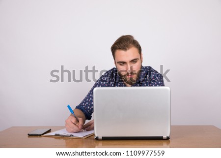 Young busy man sitting on the desk looking at the laptop working writing notes on the clipboard.