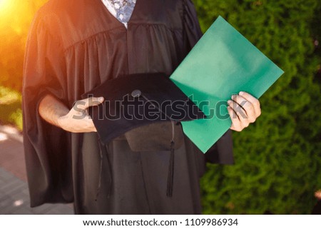 Successful graduate guy, in academic dresses, on graduation posing, holding a green folder and looking at it, farewell study, can be used for advertising,