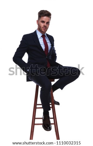 seductive businessman relaxing on wooden chair with hands in pockets, on white background, ful body picture. He is wearing a navy colored suit and a red tie