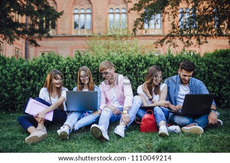 A group of students sit in a campus. Repeat course work on a laptop. In the background University.
