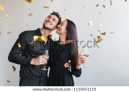 Excited man with black beard looking at confetti and embracing slim brunette woman. Indoor portrait of happy loving couple enjoying party with glasses of champagne.
