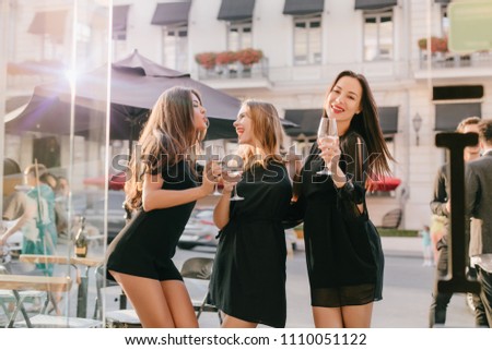 Long-haired girl in fashionable black bodysuit posing with kissing face expression looking at blonde sister. Outdoor portrait of chilling brunette lady spending time with friends and drink wine.