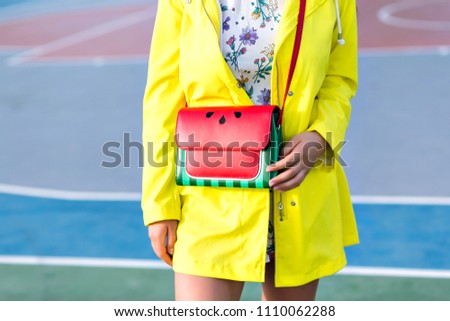 Trendy girl with red handbag posing at basketball court. Outdoor photo of female model in bright yellow coat wears stylish purse with watermelon design.
