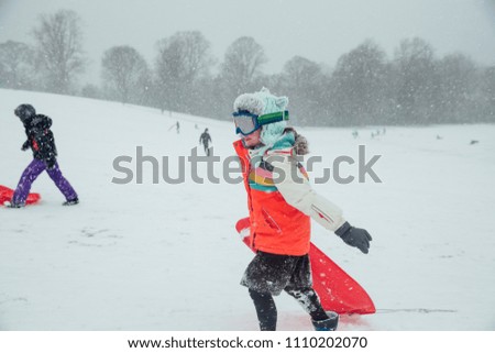 Little girl is carrying her sled back up a hill with her friends. It is snowing and she is wearing ski goggles.