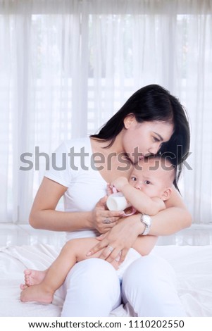 Picture of beautiful woman kissing her baby while feeding from a bottle and sitting in the bedroom