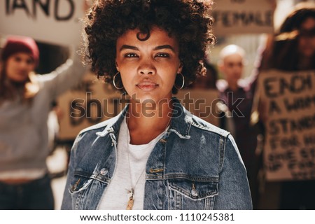 Young african woman standing outdoors with group of demonstrator in background. Woman protesting with group of activists outdoors on road. Royalty-Free Stock Photo #1110245381