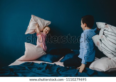 Little girl in pink t-shirt and boy in blue shirt fight with pillows on the bed