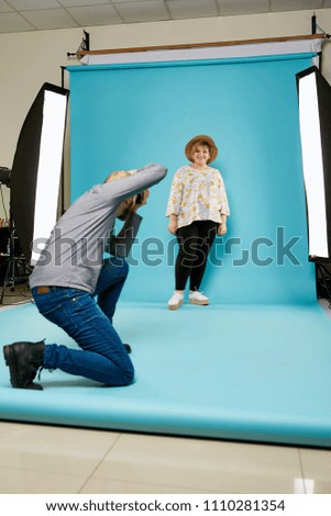 A professional photographer taking pictures of the beautiful chubby woman in the photo studio. The confident cheerful lady standing, posing over the blue background, smiling. Body positive concept.