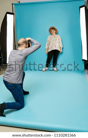 A professional photographer taking pictures of the beautiful plus-size model in the photo studio. The confident cheerful lady standing, posing over the blue background, smiling. Body positive concept.
