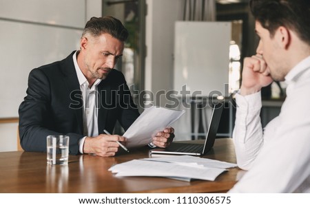 Business, career and placement concept - caucasian businessman negotiating with male candidate about work while reading his resume during job interview in office Royalty-Free Stock Photo #1110306575